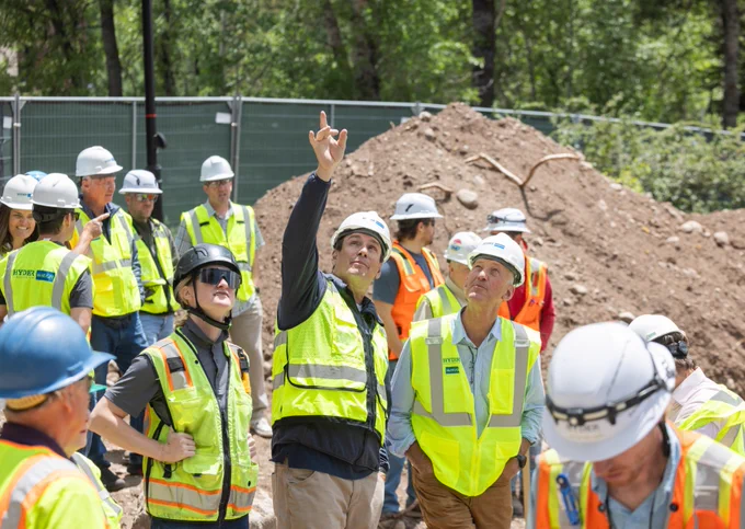 Vail Town Council and others in safety gear attentively watching a colleague pointing upwards at a construction site.