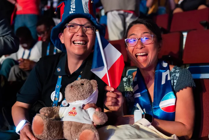 Two spectators smiling and holding a flag at the 2025 Rotary International Convention, one wearing a hat with "France" written on it and holding a End Polio Now bear, while the other is draped in a flag.