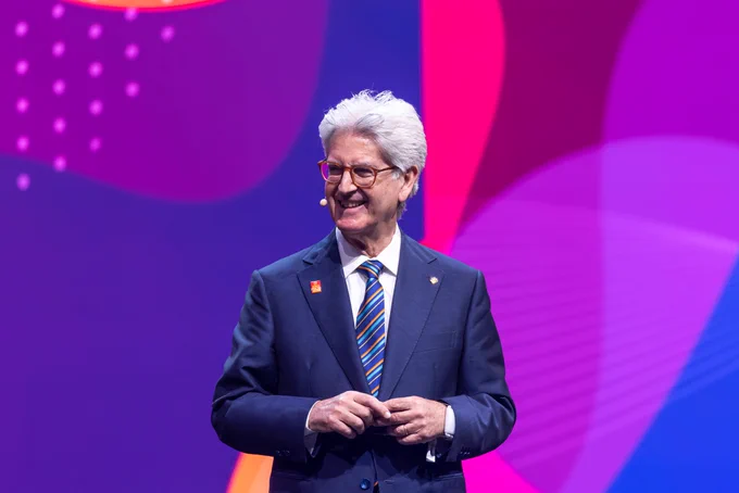 Rotary President-elect Francesco Arezzo standing on stage at the 2025 Convention, smiling with a microphone, dressed in a suit with an End Polio Now lapel pin. Colorful graphic background on stage.