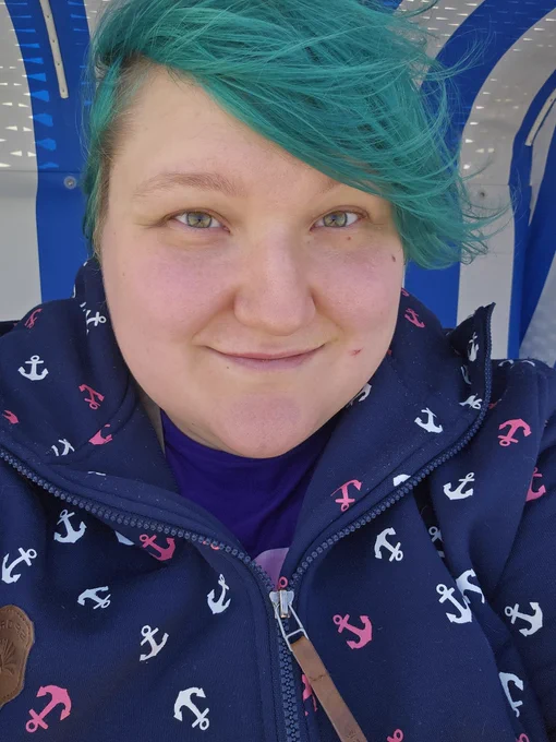 Selfie von mir in einer dunkelblauen Ankerjacke mit türkisen Haaren und einem blau weiß gestreiften Strandkorb im Hintergrund. 