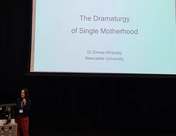Dr Emma Whipday stands behind a podium before a large screen. The title slide of her presentation reading 'the dramaturgy of single motherhood' is projected onto it 