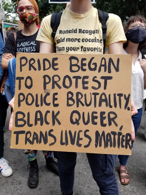 A person holds up a cardboard sign that says "PRIDE BEGAN TO PROTEST POLICE BRUTALITY. BLACK QUEER, TRANS LIVES MATTER."