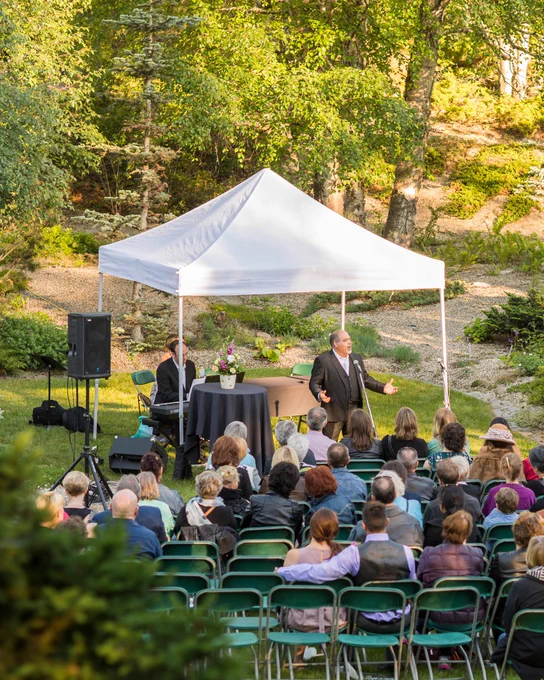 A male opera singer performs under a white canopy tent in a forested garden setting. Audience members are seated in green chairs, attentively watching the performance. A pianist sits at an electric keyboard beside a small table with floral arrangements.
