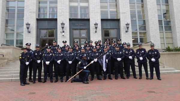 A group of police officers in uniform posing on the steps of Elle Hall, with one officer holding a flag at the front.