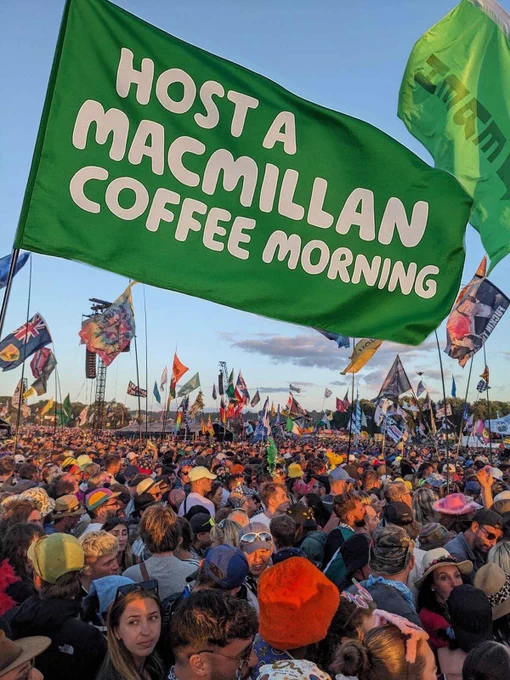 A crowd of people at Glastonbury festival with a large green flag waving above reading ‘Host a Macmillan Coffee Morning’