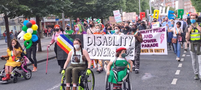 Image shows colourfully dressed participants at the Disability Pride and Power Parade 2024. At the front are two teenage girls in wheelchairs, one in a red beret and green dress, and to the left a woman in a yellow dress speaking into a mic. Behind her is a colourful set of balloons held aloft in disability bright flag colours, and more are at the back which spell POWER. In the centre participants are holding colourful banners which read “Disability Power   Pride” and “Disability Pride and Power Parade: Power in Unity” 
