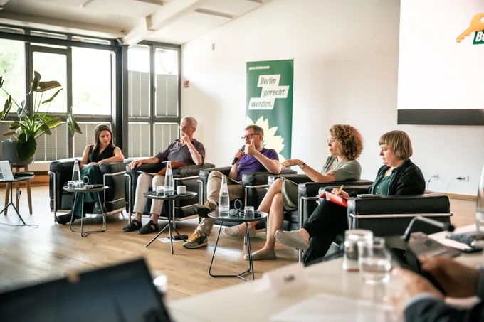 Mareile Dedekind, Rainer Tietzsch, Werner Graf, Claudia Leistner und Katrin Schmidberger sitzen in schwarzen Sesseln auf einem Podium in einem hellen Veranstaltungsraum. Sie diskutieren, machen sich Notizen oder hören aufmerksam zu. Eine Person spricht gerade ins Mikrofon. Im Hintergrund ist ein grünes Banner mit der Aufschrift „Berlin gerecht werden.“ und dem Logo der Berliner Grünen zu sehen. Auf den Tischen vor den Sprecher*innen stehen Wasserkaraffen und Gläser.