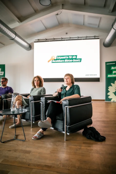 Werner Graf, Claudia Leistner und Katrin Schmidberger sitzen auf einem Podium in schwarzen Sesseln. Katrin Schmidberger spricht mit einem Mikrofon und hält Unterlagen in der Hand. Im Hintergrund ist auf einer großen Leinwand der Schriftzug „Eckpunkte für ein Bezahlbare-Mieten-Gesetz“ mit einem Schlüssel-Symbol zu sehen. Der Raum ist hell und modern gestaltet, mit Holzfußboden und sichtbaren Lüftungsrohren.