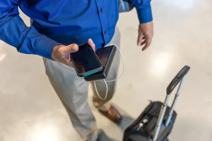 A person in a blue shirt and dark pants with a suitcase, holding a smartphone connected to a power bank.
