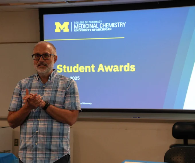A person standing in front of a presentation screen displaying "Student Awards 2025" at the University of Michigan College of Pharmacy, Medicinal Chemistry.