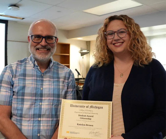 Two people smiling, holding a 'University of Michigan' award certificate titled 'Student Award for Citizenship'. Both stand in a room that appears to be an office or classroom setting.