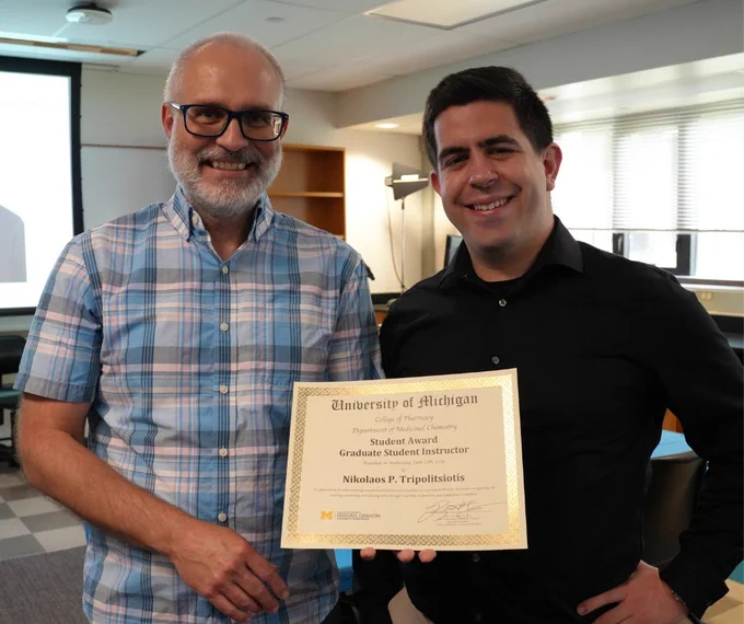 Two individuals smiling at the camera, one holding a University of Michigan Student Award certificate for Graduate Student Instructor named Nikolaos P. Triantosilits.