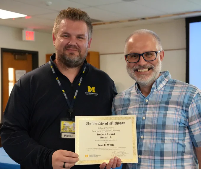 Two people smiling and holding a University of Michigan award certificate for Outstanding Student Research.