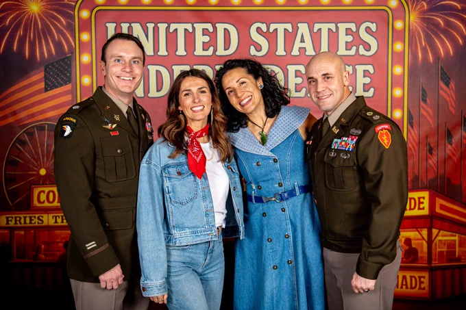 Two U.S. military officers and two guests in themed denim and red bandanas pose in front of a vibrant State Fair-style “United States Independence” backdrop.