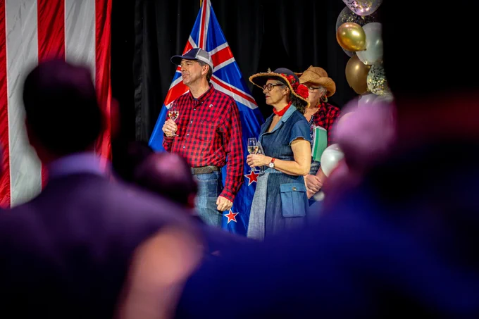 Guests in State Fair-inspired attire stand for the toast, with American and New Zealand flags in the background.