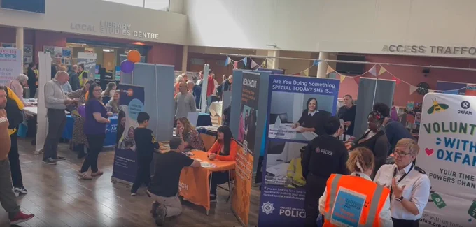 Wide view of the Volunteer Fair. More tables, stalls, and banners can be seen, filling the large room. Many people are visiting the fair.