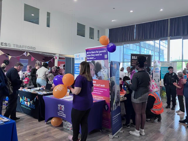 The Volunteer Fair. Stalls and tables in a large room, banners stand behind the stalls, people mill about looking at what's on offer. There are quite a few orange and blue balloons.