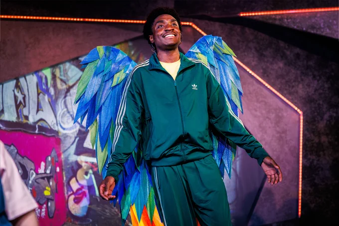 An actor on stage, wearing a tracksuit and colourful wings.
