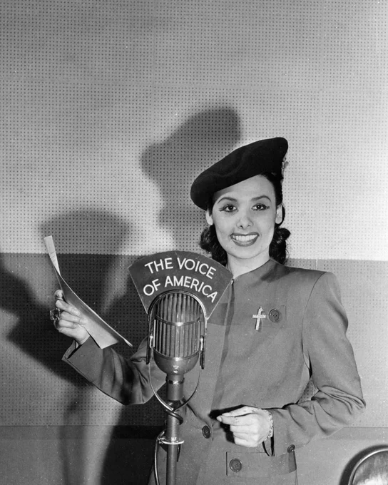 Lena Horne, smiling, stands behind a vintage microphone labeled 'The Voice of America' while holding a paper in her right hand.