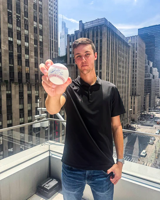 Jacob Misiorowski holds up a baseball that he has signed. He is wearing a black polo shirt and blue jeans.