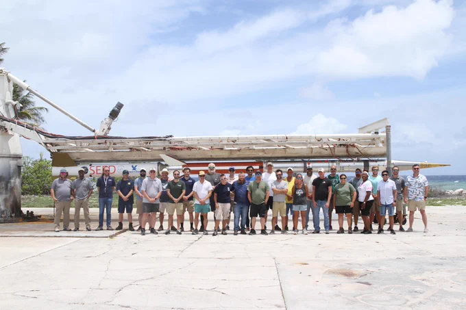 A large group of people stand in a row on a concrete surface outdoors, with a sounding rocket mounted on the launch rail behind them and the ocean visible in the background under a partly cloudy sky.
