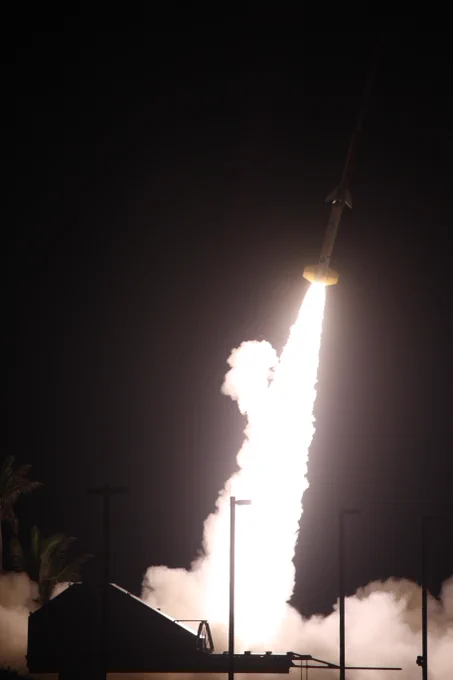 A rocket launches into the night sky, surrounded by clouds of smoke and glowing flames at its base, illuminating the dark background.