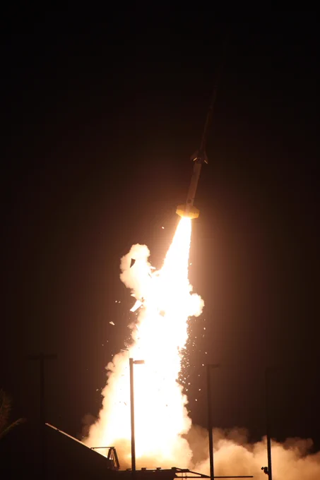 A bright rocket launches into the night sky, surrounded by clouds of smoke and glowing flames at its base, illuminating the dark background.