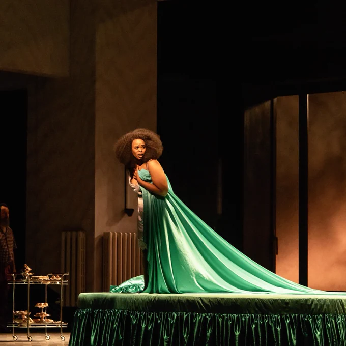 A performer onstage wearing an elegant green gown with a long train, standing in a dramatic pose, set against a minimalist theatrical backdrop.