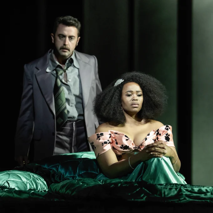 Two opera performers on stage, one seated on a bed in a floral dress and the other standing beside in a suit.