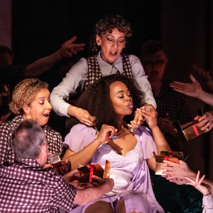 A dramatic stage scene with multiple performers engaging vividly around a central figure in a white dress, who is receiving gifts from others. The setting suggests an intense and emotional moment in a theatrical production.