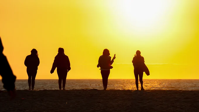 Four silhouettes walking on a beach at sunset, with a bright sun low on the horizon.