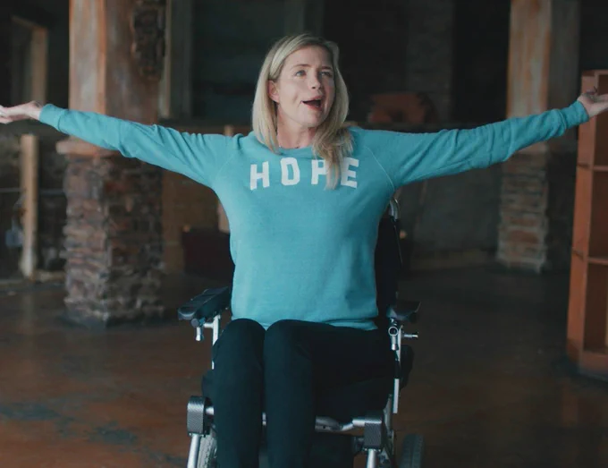 A woman sits in a wheelchair in an empty building with her arms spread wide. She wears a blue shirt with the words “Hope” across the chest.