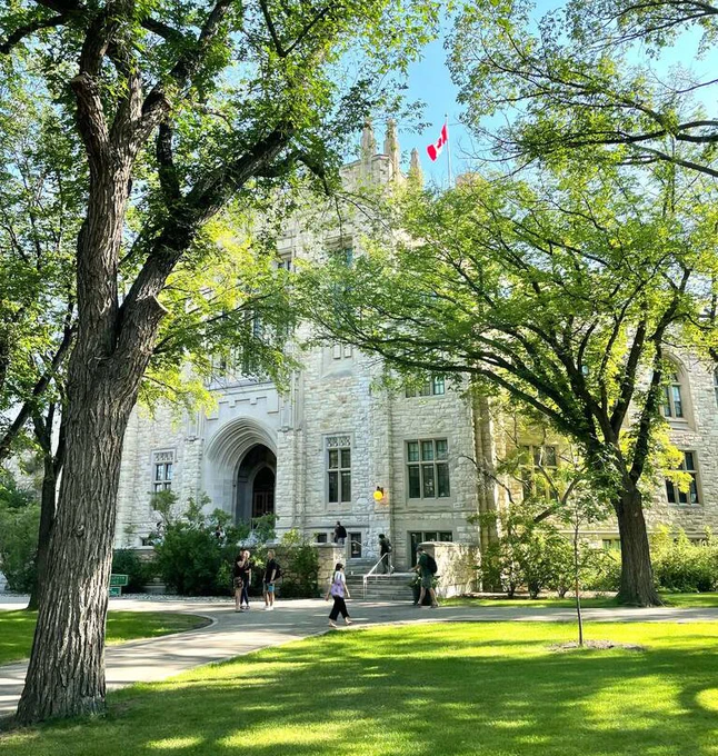 People walking near the entrance of the Thorvaldson Building, surrounded by lush green trees on a sunny day.

