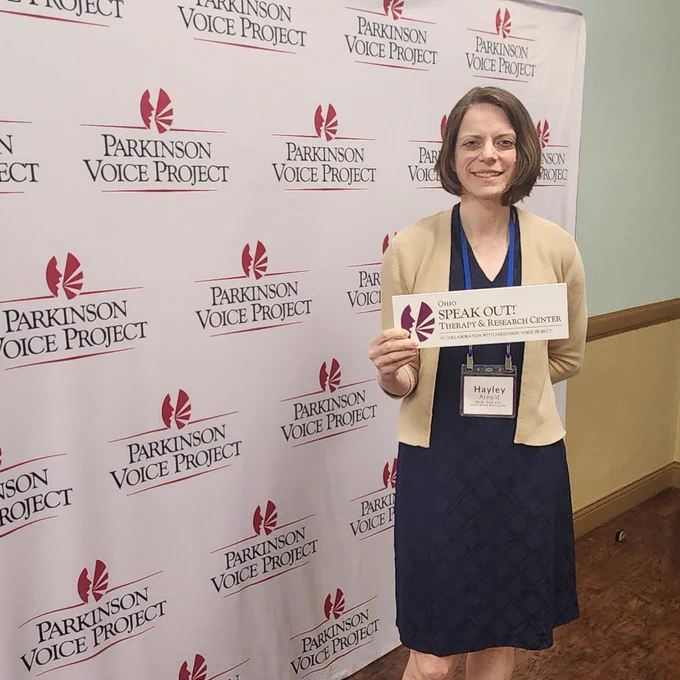 Kent State faculty member Hayley Arnold standing in front of a Parkinson Voice Project backdrop as she holds a sign that reads 'Ohio SPEAK OUT! Therapy and Research Center.'