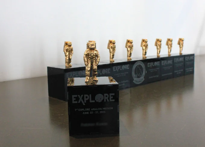 Golden astronaut figurines mounted on black bases labeled 'EXPLORE' for the 1st Explore Analog Mission, lined up on a reflective surface.
