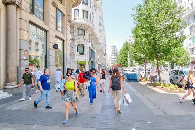 People in casual summer attire walk and sit along the wide sidewalks of Gran Vía, Madrid’s central avenue lined with retail storefronts, tall buildings, shade trees, lighting fixtures, and wooden benches.