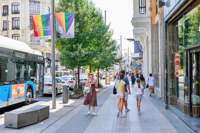 People in casual summer attire walk and sit along the wide sidewalks of Gran Vía, Madrid’s central avenue lined with retail storefronts, tall buildings, shade trees, lighting fixtures, and wooden benches.