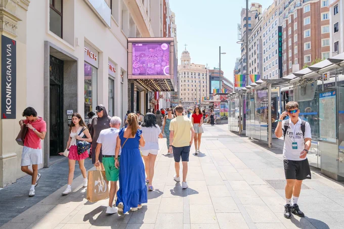 People in casual summer attire walk and sit along the wide sidewalks of Gran Vía, Madrid’s central avenue lined with retail storefronts, tall buildings, shade trees, lighting fixtures, and wooden benches.