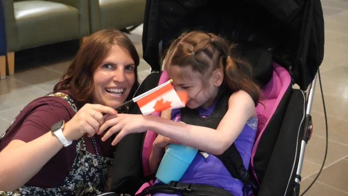 Two individuals smiling, one holding a small Canadian flag while seated beside a younger individual in a stroller. 