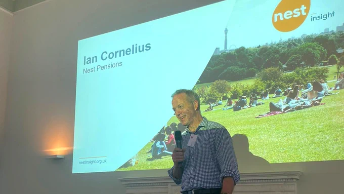 Ian Cornelius of Nest Pensions stands speaking into a microphone in front of a projected slide at a Nest Insight event. The slide displays his name and organisation alongside an image of people sitting on grass in a park, with the BT Tower visible in the background. The Nest Insight logo is in the top right corner.