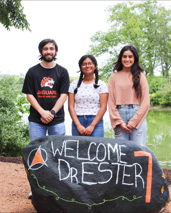 Three individuals standing behind a large rock painted with the text "Welcome Dr. Ester" at a park.