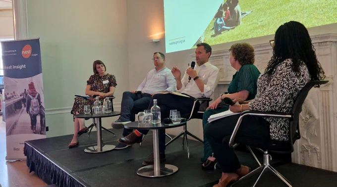 Five panel speakers are seated on a stage at a Nest Insight event. One man is speaking into a microphone while others listen. A screen behind them displays the Nest Insight website and a photo of people sitting on grass. A branded Nest Insight banner stands to the left. Water bottles and glasses are on small tables in front of the speakers.