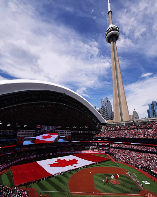 High-angle photo from Rogers Centre in Toronto shows a full Blue Jays stadium with a gigantic Canada flag unfurled across the outfield. The CN Tower looms in the background beneath a cloudy blue sky. (Photo: Michael Chisholm/Major League Baseball)