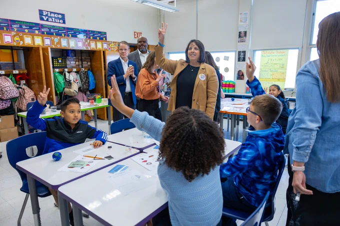 A classroom filled with elementary school students and several adult visitors. Students sit at tables raising their hands enthusiastically, while Lt. Governor Kim Driscoll, wearing a tan blazer and a "STEM Week" button, also raises her hand in a peace sign. Other adults, including a man clapping and another standing beside him, observe and engage with the students. The classroom is colorful, with educational posters and backpacks hanging in cubbies.