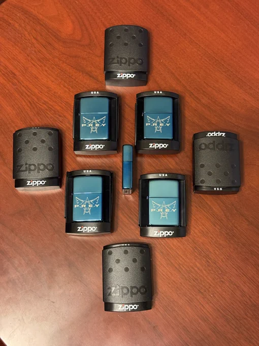A collection of PREY (2006)-branded Zippo lighters arranged neatly in a circular formation on a wooden surface. Six of the lighters display the blue PREY logo with the Talon symbol, surrounded by black Zippo hand warmers and cases. A small blue USB stick is positioned in the center of the layout.