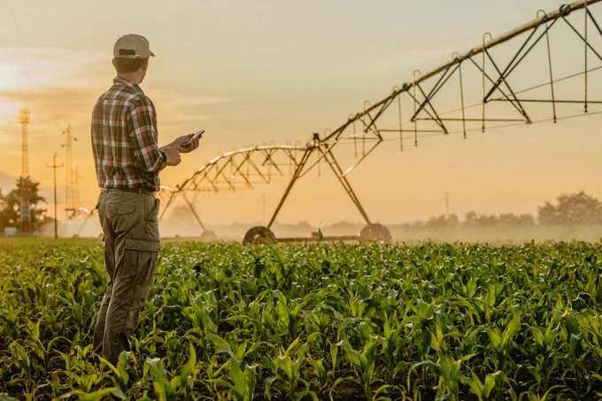 A farmer standing in a field at sunrise, examining a smart phone with a large irrigation system in the background.
