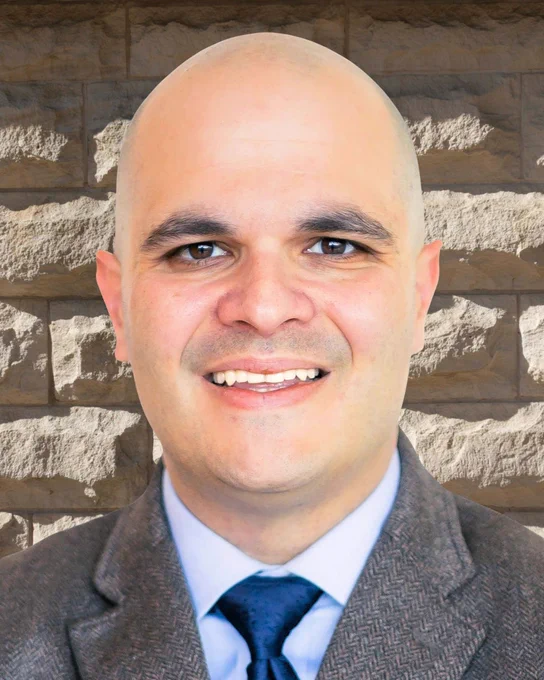 A professional headshot of a smiling Dr. Varisco wearing a gray suit and navy tie, standing against a brick wall background.