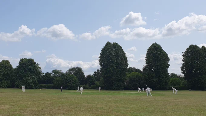 View from boundary shows fielders, bowler and batter waiting to hit ball  as umpire looks on