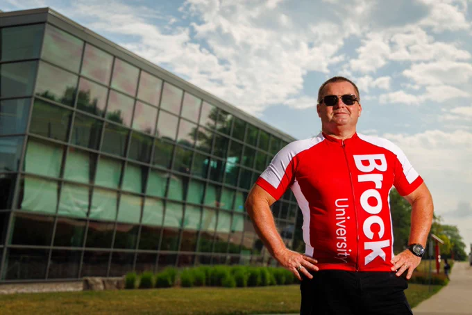 A man wearing sunglasses and a red-and-white Brock University cycling jersey stands confidently with hands on hips in front of a modern glass building under a partly cloudy sky.
