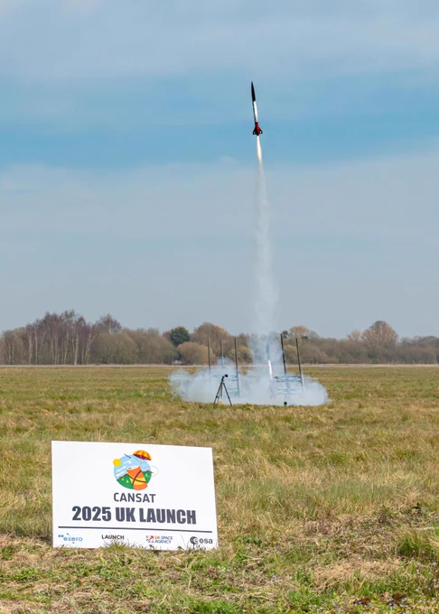 A rocket launching at the 2025 UK launch event of CanSat, with a launchpad visible in a grassy field under clear skies. A sign featuring logos from sponsors including Airbus, UK Space, and ESA is prominently displayed in the foreground.